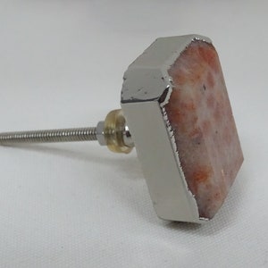 May include: A square-shaped cabinet knob with a silver metal frame and a light orange and white sunstone inlay. The knob has a silver metal screw.