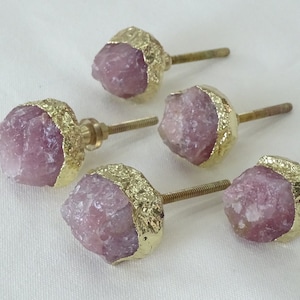 Könnte beinhalten: Vier rosa Kristall-Schrankknöpfe mit goldenen Akzenten. Jeder Knopf hat eine einzigartige Form und ist an einer goldenen Schraube befestigt.