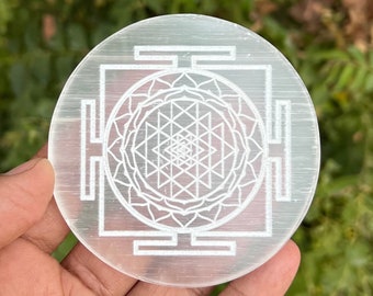 Puede incluir: Un cristal de selenita blanco y redondo con un Sri Yantra grabado en su superficie. El cristal tiene un diámetro de aproximadamente 5 cm.