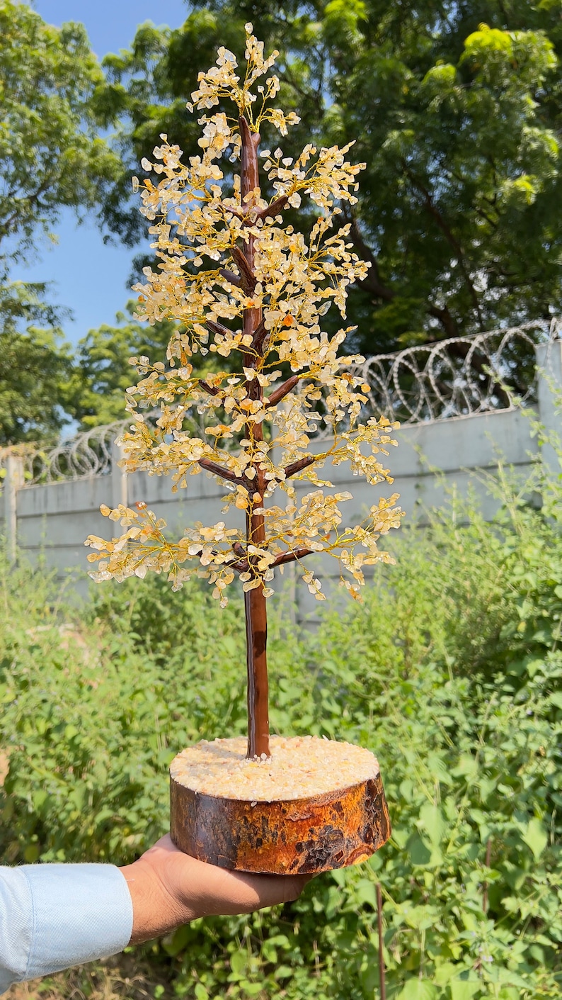 May include: A crystal tree made of clear and amber colored stones, with a brown wooden base. The tree is about 12 inches tall and is displayed on a hand.