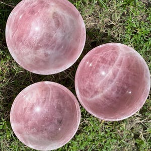 May include: Three polished rose quartz spheres, each displaying a soft pink hue with natural veining. The spheres are arranged on a bed of green grass, showcasing their smooth, reflective surfaces. These decorative spheres are ideal for home decor.