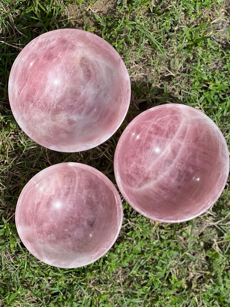 May include: Three polished rose quartz spheres, each displaying a soft pink hue with natural veining. The spheres are arranged on a bed of green grass, showcasing their smooth, reflective surfaces. These decorative spheres are ideal for home decor.