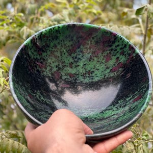 May include: A green and black bowl with pink and white flecks. The bowl is made of polished stone and has a smooth, rounded surface.