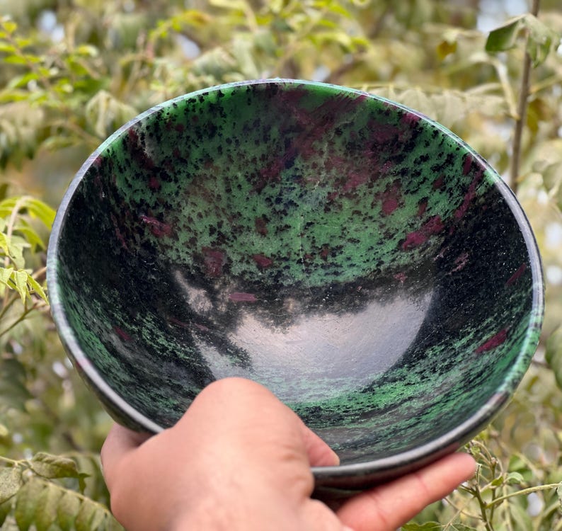 May include: A green and black bowl with pink and white flecks. The bowl is made of polished stone and has a smooth, rounded surface.