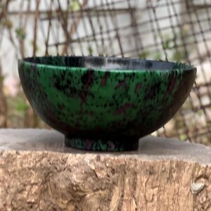 May include: A green and black speckled bowl with a rounded shape. The bowl is sitting on a tree stump.
