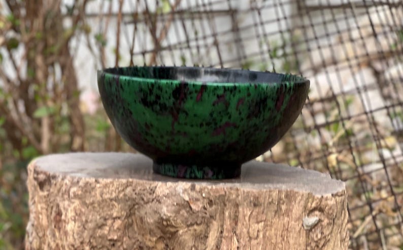 May include: A green and black speckled bowl with a rounded shape. The bowl is sitting on a tree stump.