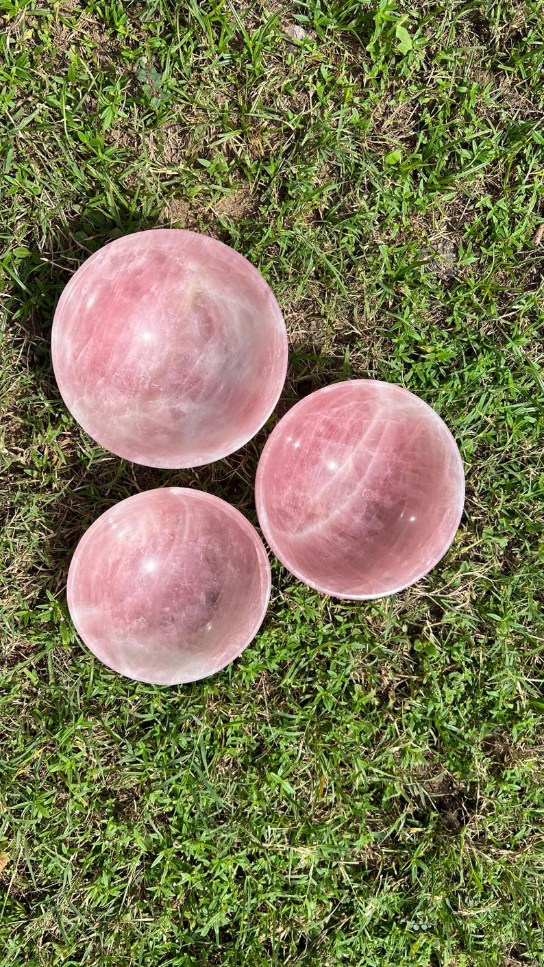 May include: Three polished rose quartz spheres, each with a soft pink hue and natural veining. The spheres are displayed on a bed of green grass, creating a natural and earthy aesthetic. These spheres are ideal for home decor or crystal healing.