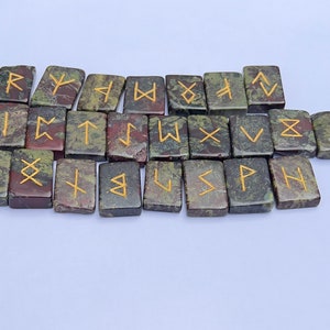以下が含まれることがあります： 金色の文字が書かれた、緑色、茶色、黒色の石のルーン24個のセット。各ルーンは、ユニークなシンボルが刻まれた小さな長方形の石です。