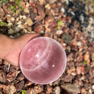 May include: A polished rose quartz sphere, showcasing a soft pink hue with subtle internal veining. The sphere is smooth and round, reflecting light. This decorative crystal ball is a natural stone, perfect for home decor or spiritual practices.