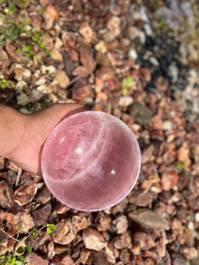 May include: A polished rose quartz sphere, showcasing a soft pink hue with subtle internal veining. The sphere is smooth and round, reflecting light. This decorative crystal ball is a natural stone, perfect for home decor or spiritual practices.