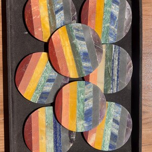 May include: A set of six circular coasters arranged in a black tray. Each coaster features vertical stripes of multi-coloured natural stone, including red, orange, yellow, green, blue, and purple segments, creating a vibrant, layered appearance.