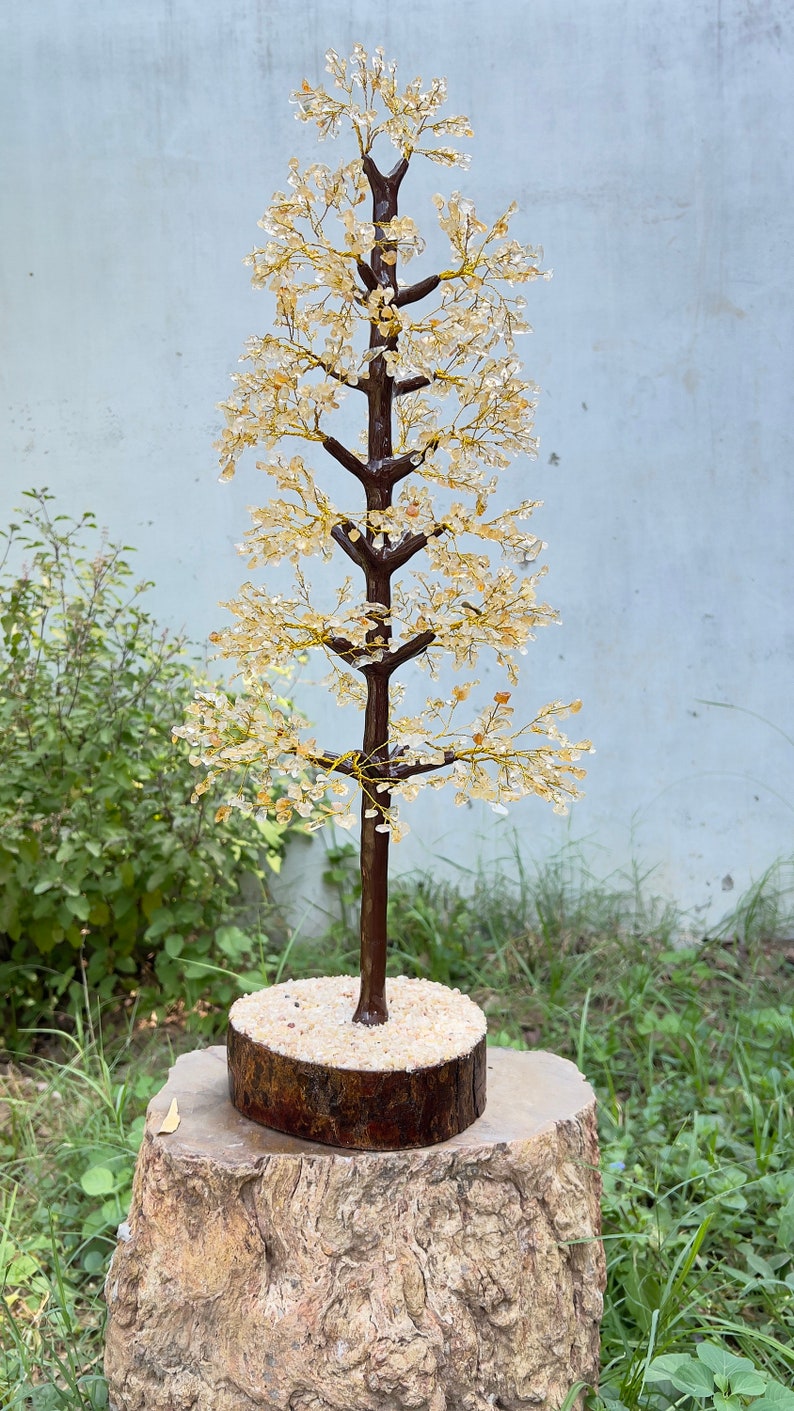 May include: A crystal tree made of clear and amber-colored stones, with a brown wooden base and a sand-filled pot. The tree is on a weathered tree stump.