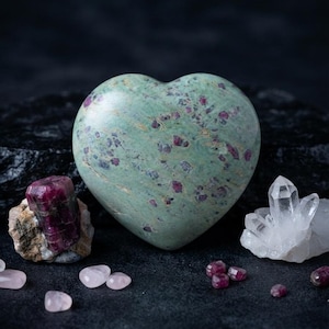May include: A heart-shaped polished stone featuring a light green base with scattered deep pink and magenta inclusions. The stone is surrounded by a raw dark pink crystal, a cluster of clear quartz points, and several small loose pink and red stones.
