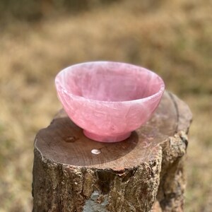 May include: A pink rose quartz bowl with a rounded shape and a small, circular base. The bowl is set on a weathered, brown tree stump against a blurred, natural background. The bowl's color is a soft, translucent pink.