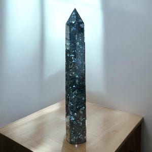 Pode incluir: Uma grande ponta de cristal facetado, transparente, com inclusões pretas e prateadas. O cristal está sobre uma superfície de madeira clara.
