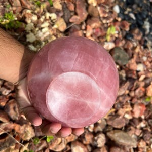 May include: A polished rose quartz sphere, showcasing a soft pink hue with natural veining. The sphere has a flat base and is held in a hand, set against a background of small rocks and greenery. This decorative item is ideal for home decor.