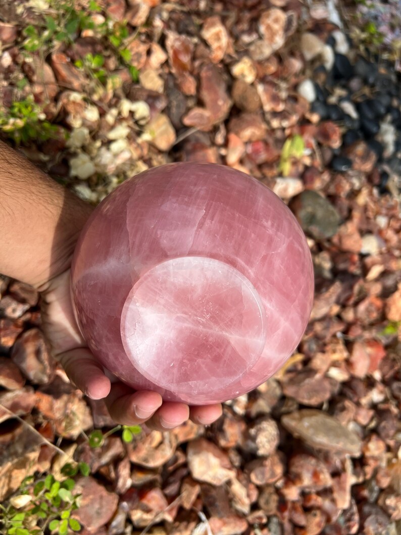 May include: A polished rose quartz sphere, showcasing a soft pink hue with natural veining. The sphere has a flat base and is held in a hand, set against a background of small rocks and greenery. This decorative item is ideal for home decor.