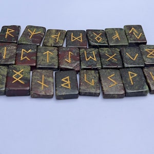 以下が含まれることがあります： 金色の文字が書かれた緑と赤の石のルーン24個のセット。ルーンは2列に並べられており、上の列は下の列から少しずれています。ルーンはすべて長方形で、滑らかで磨かれた表面をしています。