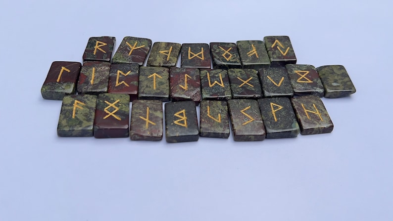 以下が含まれることがあります： 金色の文字が書かれた緑と赤の石のルーン24個のセット。ルーンは2列に並べられており、上の列は下の列から少しずれています。ルーンはすべて長方形で、滑らかで磨かれた表面をしています。