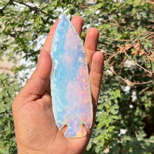 Puede incluir: Una punta de flecha de ópalo iridiscente con un esquema de color azul claro y rosa. La punta de flecha se sostiene en una mano contra un fondo verde de hojas.
