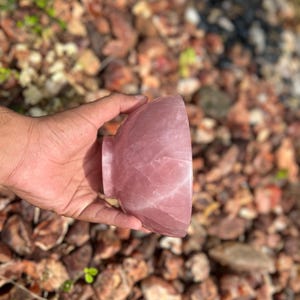 May include: A polished, rose-colored quartz bowl with a rounded shape and a small, circular base. The bowl is held in a hand against a background of small, brown and tan rocks.