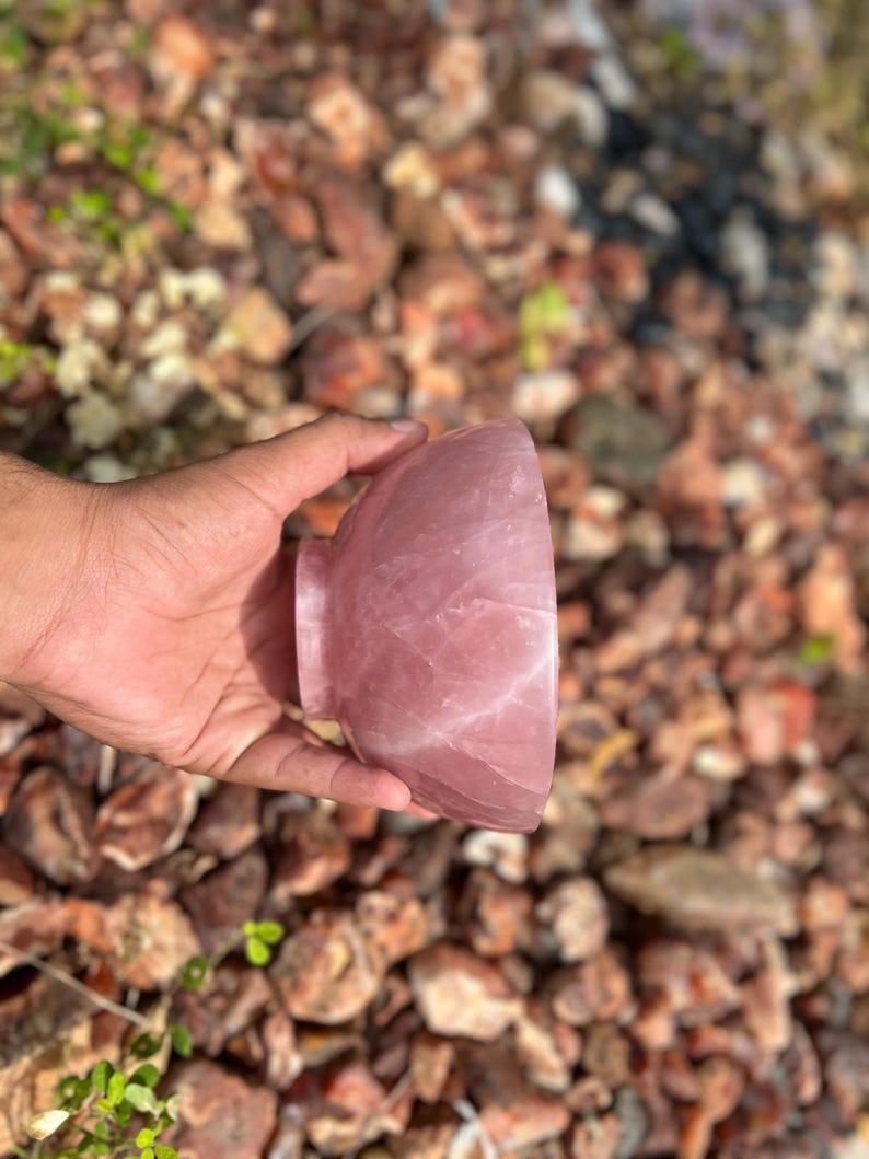 May include: A polished, rose-colored quartz bowl with a rounded shape and a small, circular base. The bowl is held in a hand against a background of small, brown and tan rocks.