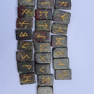 以下が含まれることがあります： 金色の文字が刻まれた緑と赤の石のルーン24個のセット。ルーンは白い背景に半円形に配置されています。