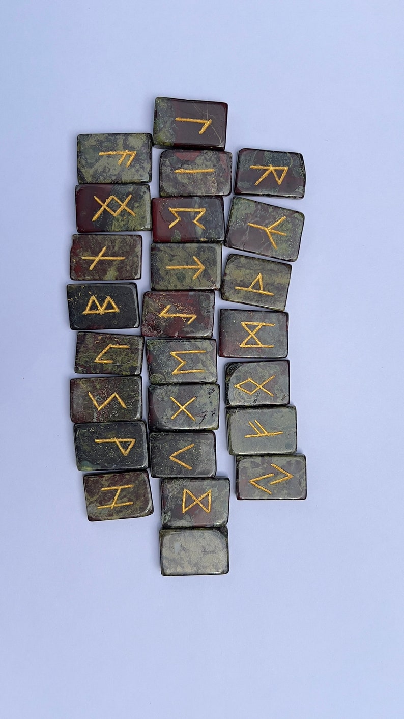 以下が含まれることがあります： 金色の文字が刻まれた緑と赤の石のルーン24個のセット。ルーンは白い背景に半円形に配置されています。