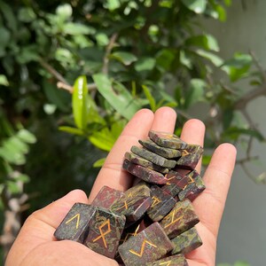 以下が含まれることがあります： 手の中に緑、赤、金の石のルーンのセットが置かれている。ルーンは、金色のシンボルが刻まれた小さな長方形の石の形をしている。