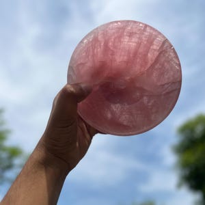 May include: A large, round, polished rose quartz crystal bowl, held up against a blue sky with some clouds. The bowl is a soft pink color and appears translucent, with visible internal patterns.