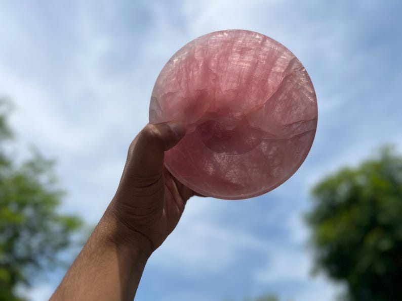 May include: A large, round, polished rose quartz crystal bowl, held up against a blue sky with some clouds. The bowl is a soft pink color and appears translucent, with visible internal patterns.