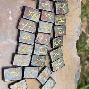 以下が含まれることがあります： 金色の文字が刻まれた緑と赤の石のルーン24個のセット。ルーンは茶色の表面にランダムなパターンで配置されています。
