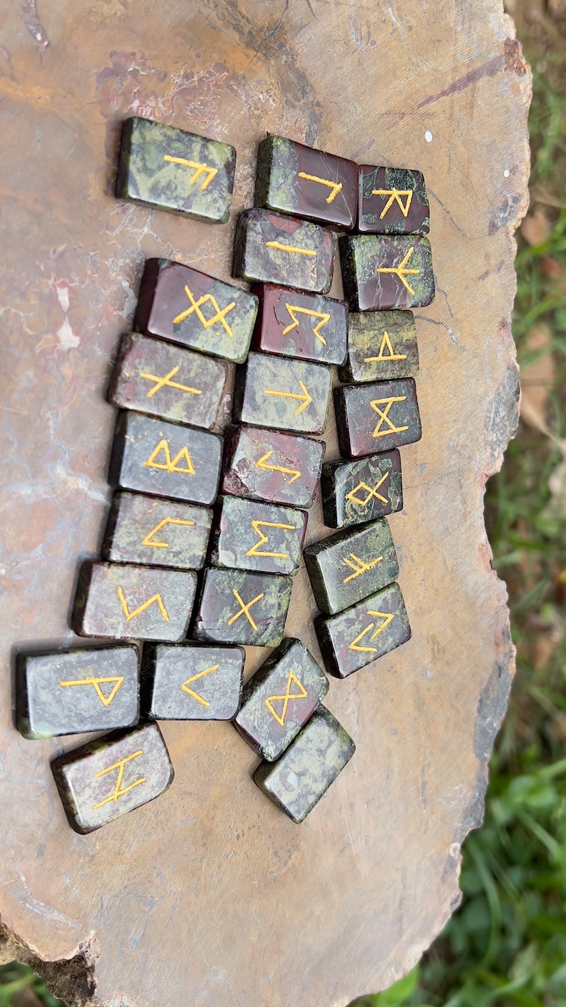 以下が含まれることがあります： 金色の文字が刻まれた緑と赤の石のルーン24個のセット。ルーンは茶色の表面にランダムなパターンで配置されています。