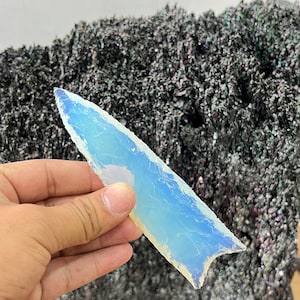 Puede incluir: Una punta de flecha de ópalo de color azul claro iridiscente, sostenida en una mano, sobre un fondo gris oscuro.