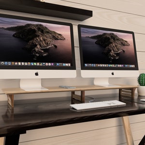 Puede incluir: Dos ordenadores iMac plateados con biseles negros est&aacute;n sobre un soporte de monitor de madera. El soporte est&aacute; sobre un escritorio marr&oacute;n oscuro con un teclado blanco y una alfombrilla de rat&oacute;n negra. Un peque&ntilde;o cactus est&aacute; sobre el escritorio a la derecha de los ordenadores.