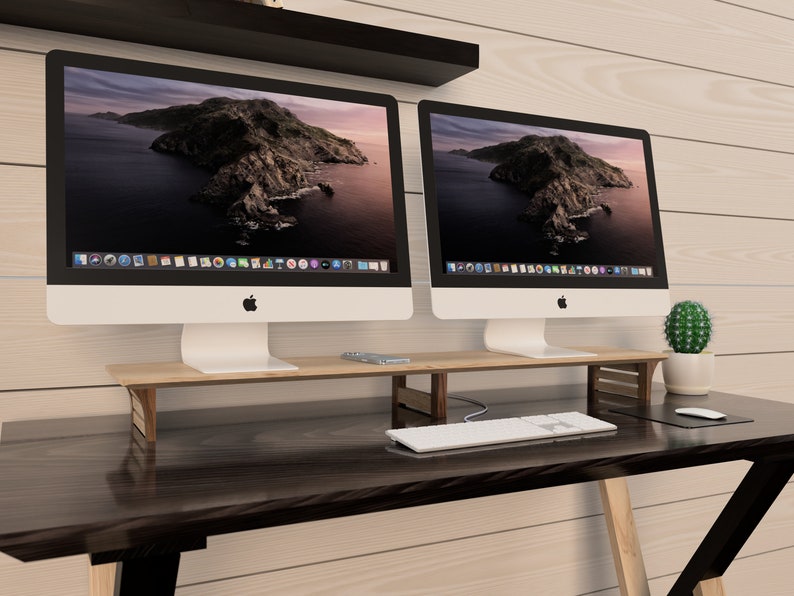 Puede incluir: Dos ordenadores iMac plateados con biseles negros est&aacute;n sobre un soporte de monitor de madera. El soporte est&aacute; sobre un escritorio marr&oacute;n oscuro con un teclado blanco y una alfombrilla de rat&oacute;n negra. Un peque&ntilde;o cactus est&aacute; sobre el escritorio a la derecha de los ordenadores.