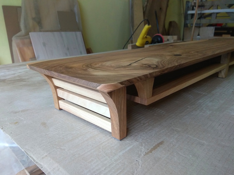Puede incluir: Un soporte de TV de madera con un acabado de madera natural y un dise&ntilde;o curvo. El soporte tiene tres estantes y una superficie superior ancha.