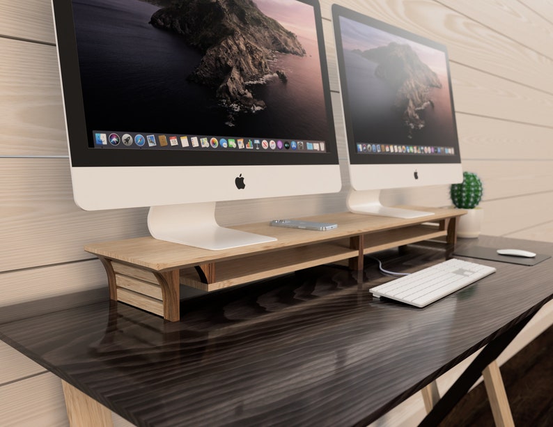 Puede incluir: Dos ordenadores iMac blancos con un soporte de monitor de madera sobre un escritorio de madera oscura. El soporte tiene un estante para guardar objetos.