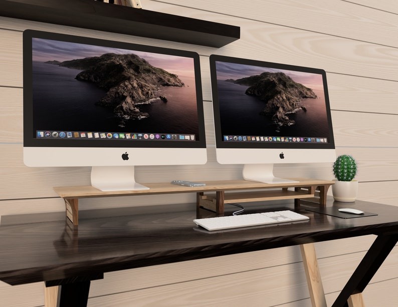Puede incluir: Dos ordenadores iMac blancos con biseles negros est&aacute;n sobre un soporte de monitor de madera. Los ordenadores est&aacute;n sobre un escritorio marr&oacute;n oscuro con un teclado blanco y un rat&oacute;n blanco. Un peque&ntilde;o cactus en maceta est&aacute; sobre el escritorio a la derecha de los ordenadores.