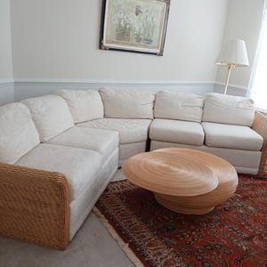 May include: A sectional sofa with cream-colored cushions and wicker armrests. A round, light brown coffee table sits on a red patterned rug. A framed floral painting hangs on the wall.