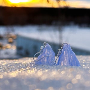 May include: Two delicate, light blue earrings with silver hooks, resting on a snowy surface. The earrings are shaped like small, frosted mountains.