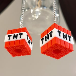 Może przedstawiać: Dwa czerwono-białe wisiorki w kształcie bloku TNT zawieszone na srebrnych łańcuszkach. Każdy sześcianowy wisiorek ma czarne litery "TNT" na białym pasku. Wisiorki mają szczegółowy, teksturowany design.