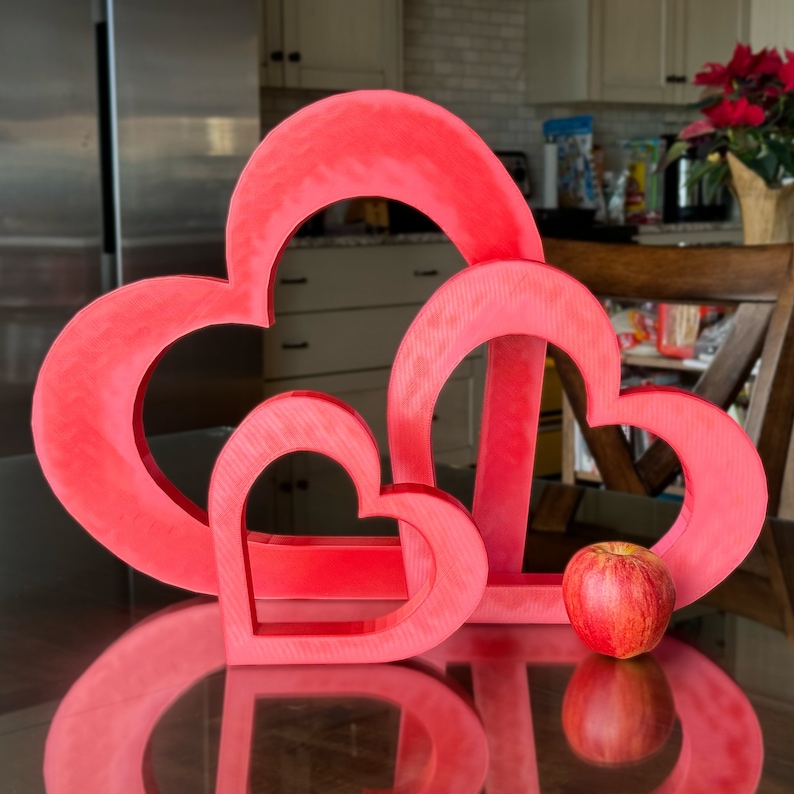 May include: Three red heart-shaped decorations, nested inside each other, with the largest heart at the back and the smallest heart in the front. A red apple sits in front of the hearts.