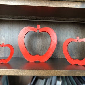 May include: Three red apple-shaped wall decorations of varying sizes on a wooden shelf.