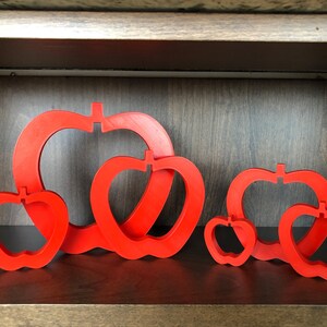 May include: A set of five red wooden apple-shaped ornaments. The ornaments are nested inside each other, with the largest at the back and the smallest at the front.