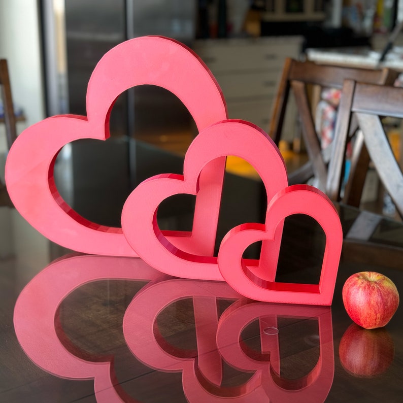 May include: Three red heart-shaped decorations of varying sizes, arranged in a descending order of size, on a wooden surface. A red apple sits to the right of the hearts.