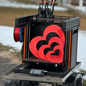 May include: A black 3D printer with a red, white and blue filament spool. The printer is printing three red hearts, one inside the other. The printer is on a metal platform with wheels.