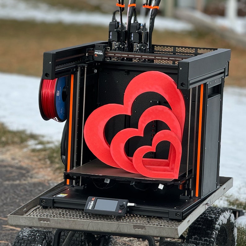 May include: A black 3D printer with a red, white and blue filament spool. The printer is printing three red hearts, one inside the other. The printer is on a metal platform with wheels.