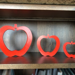 May include: Three red apple-shaped cutouts of varying sizes are displayed on a wooden shelf. The largest cutout is on the left, the medium-sized cutout is in the centre, and the smallest cutout is on the right.