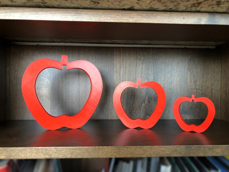 May include: Three red apple-shaped cutouts of varying sizes are displayed on a wooden shelf. The largest cutout is on the left, the medium-sized cutout is in the centre, and the smallest cutout is on the right.